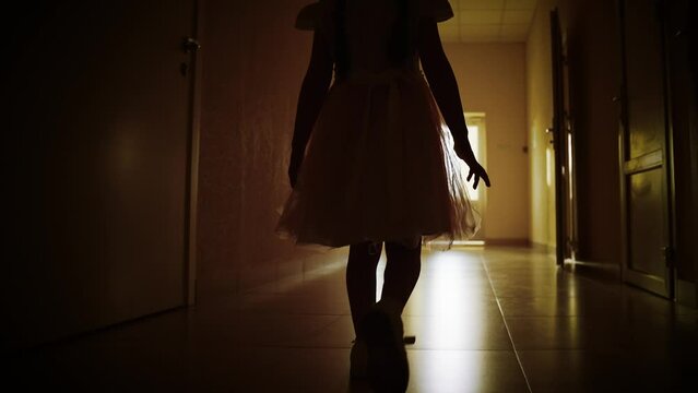 Girl Child In Dark Empty Corridor. Little Girl Looks Out Of Window, Rear View. Lonely Silhouette Of Child Running Down Dark Corridor Towards Door And Light. Primary School Looking To Future Of Child