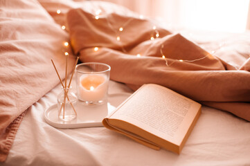 Burning candle with aroma sticks in bottle on tray with open book in bed over glowing Christmas...