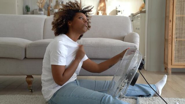 Millennial African American girl sitting on floor in living room feeling hot during extreme summer heat, using electric fan at home, young unhappy black women dealing with summertime hot flashes