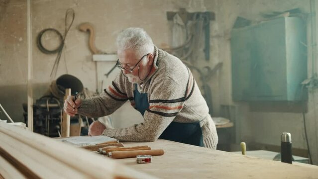 Elderly carpenter writing down notes on sheets of paper and in a book. Teamwork of an old man and a teenager in a carpentry workshop. 