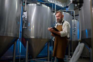 Naklejka na ściany i meble Handsome bearded man brewer inside modern beer factory around steel tanks