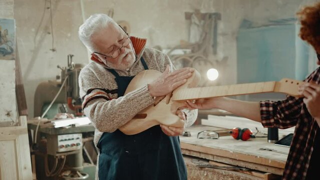 An old carpenter and his grandson look together and admire their handmade guitar at home in the workshop