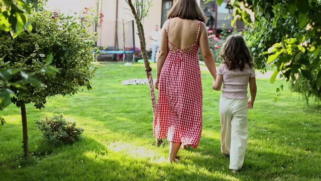 Family day, mother's day. Back view of young mom and little child daughter holds hands while walking on the backyard lawn. Idyllic family having fun and spending time outdoors on summer holiday.