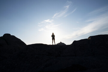 Hiking at sunset. Portrait of a young woman standing in the rocky hill with the sun hiding at her back. View of the female dark silhouette and sunny sky. 