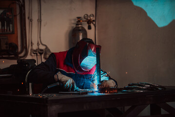 Profesional welder in protective uniform and mask welding metal pipe on the industrial table with other workers behind in the industrial workshop.