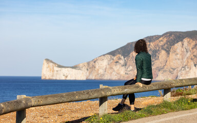 Woman Traveler sitting on the side of the highway on the Sea Coast. Sardinia, Italy. Sunny Fall Season Day.