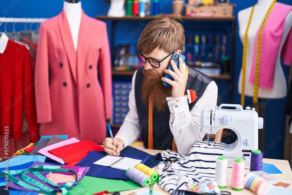 Poster young redhead man tailor talking on smartphone writing on notebook at clothing factory