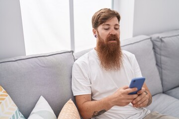Young redhead man using smartphone with serious expression at home