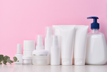 White plastic tubes, jars, and containers for cosmetic products on a pink background, advertising...