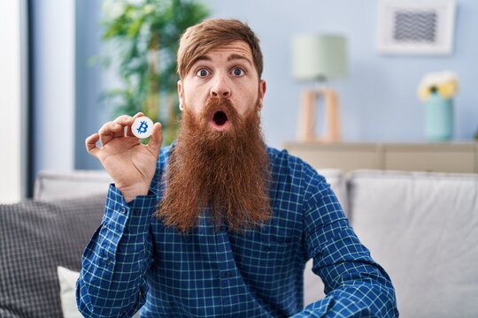 Caucasian man with long beard holding virtual currency bitcoin scared and amazed with open mouth for surprise, disbelief face