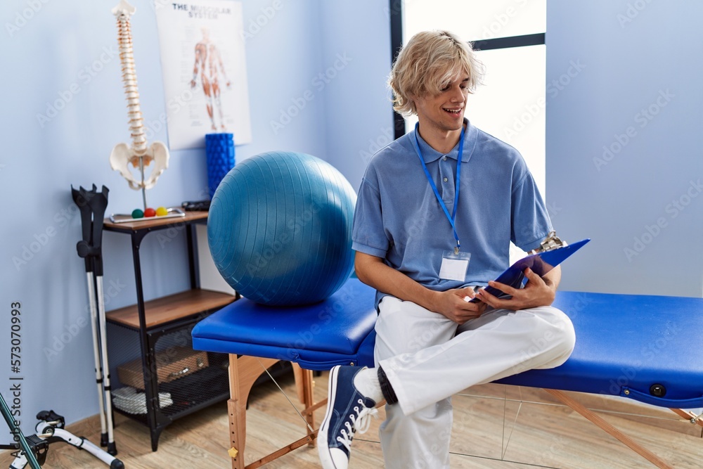 Sticker Young blond man pysiotherapist reading document sitting on massage board at rehab clinic