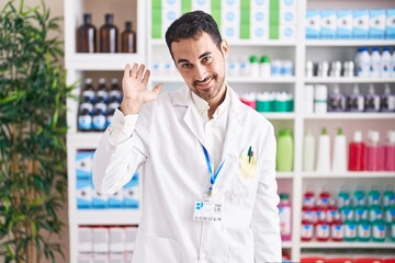 Handsome hispanic man working at pharmacy drugstore waiving saying hello happy and smiling, friendly welcome gesture