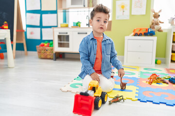Adorable hispanic toddler playing with tractor and dino toy sitting on floor at kindergarten