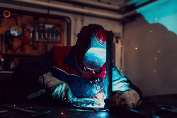 Profesional welder in protective uniform and mask welding metal pipe on the industrial table with other workers behind in the industrial workshop.