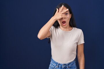 Young hispanic woman standing over blue background peeking in shock covering face and eyes with hand, looking through fingers with embarrassed expression.