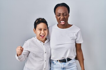 Young mother and son standing together over white background angry and mad screaming frustrated and furious, shouting with anger. rage and aggressive concept.