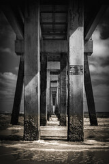 Symmetrical geometry and shapes under the Pier with poles, pillars, sunlight, ocean waves, and dramatic cloudy sky in Jacksonville Beach, Florida, USA, retro-style black and white monochromatic photo