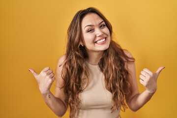 Young hispanic woman standing over yellow background success sign doing positive gesture with hand, thumbs up smiling and happy. cheerful expression and winner gesture.