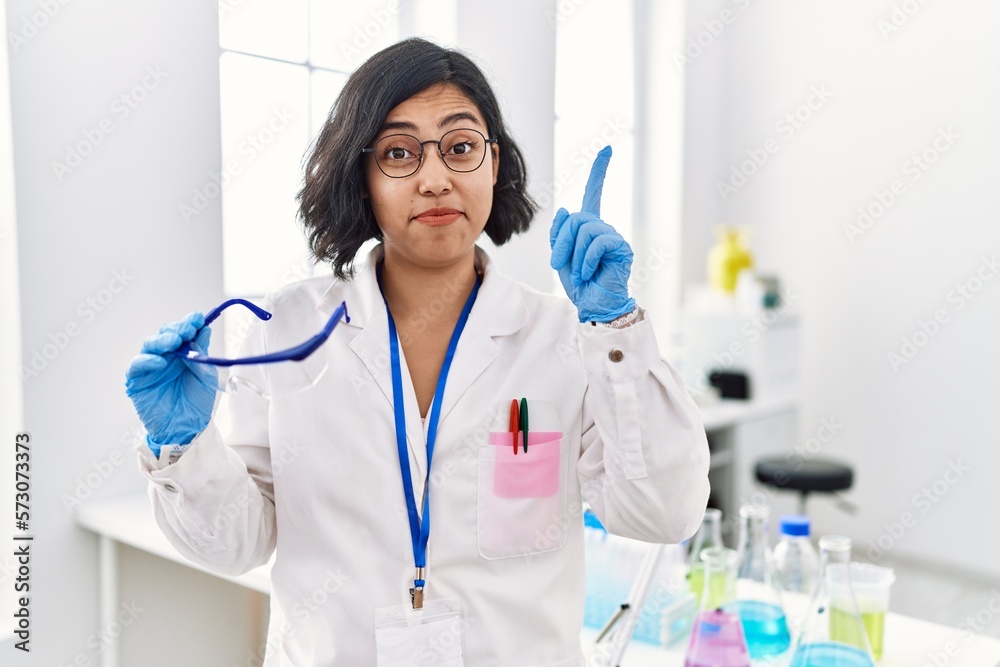 Poster Young hispanic woman working at scientist laboratory holding safety glasses smiling with an idea or question pointing finger with happy face, number one