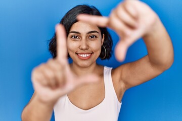 Fototapeta na wymiar Young hispanic woman standing over blue background smiling making frame with hands and fingers with happy face. creativity and photography concept.