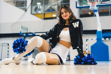 full shot of a cheerleader in a black-white uniform wearing blue pom-poms sitting on the floor....