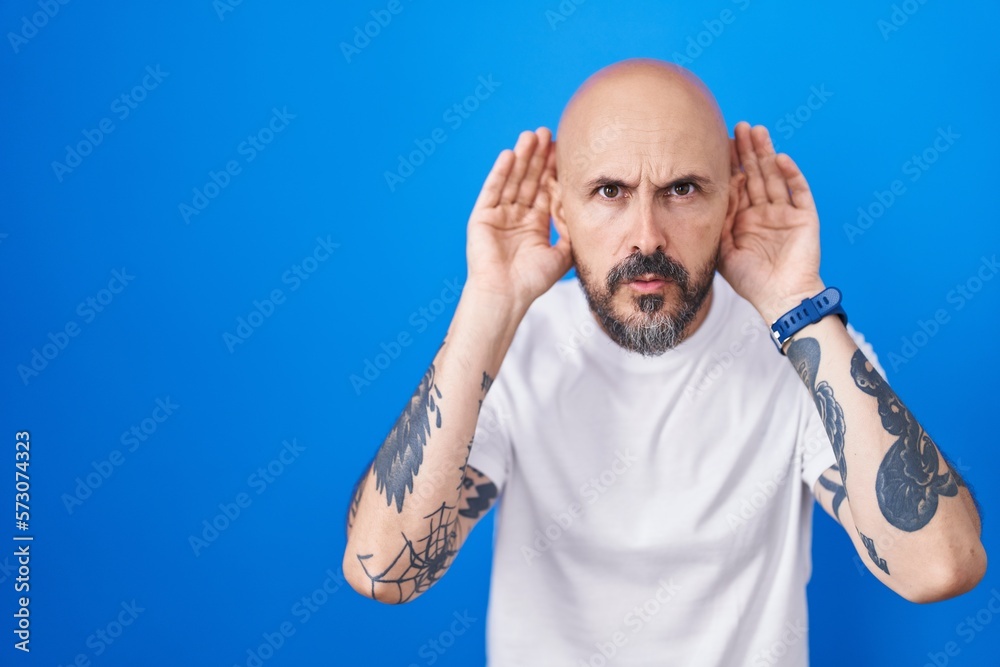 Poster hispanic man with tattoos standing over blue background trying to hear both hands on ear gesture, cu