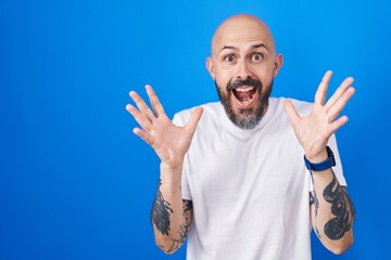 Hispanic man with tattoos standing over blue background celebrating crazy and amazed for success with arms raised and open eyes screaming excited. winner concept