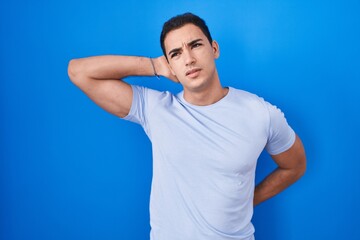 Young hispanic man standing over blue background suffering of neck ache injury, touching neck with hand, muscular pain