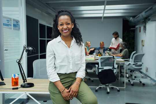 Portrait of smiling young African American professional female sitting at modern office desk. Happy mixed race business woman team leader posing for photo at corporate meeting. Concept of workplace. 
