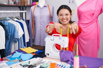 Hispanic young woman dressmaker designer using sewing machine pointing to you and the camera with fingers, smiling positive and cheerful