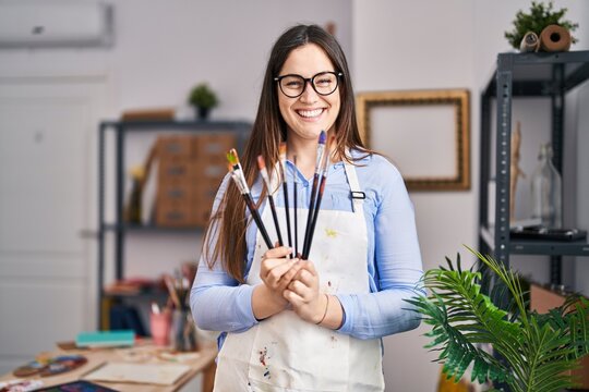 Young woman artist smiling confident holding paintbrushes at art studio