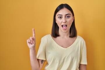 Hispanic girl wearing casual t shirt over yellow background pointing finger up with successful idea. exited and happy. number one.