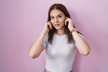 Young hispanic girl standing over pink background covering ears with fingers with annoyed expression for the noise of loud music. deaf concept.