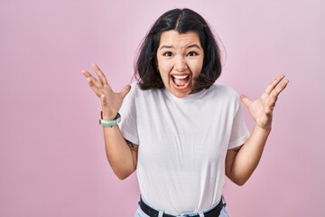 Young hispanic woman wearing casual white t shirt over pink background celebrating mad and crazy for success with arms raised and closed eyes screaming excited. winner concept