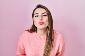 Young hispanic woman standing over pink background looking at the camera blowing a kiss on air being lovely and sexy. love expression.