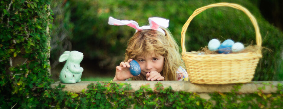 Child with rabbit ears, Easter holidays. Kid hunting Easter eggs in easters basket. Cute bunny boy, funny kids portrait. Wide photo banner for website header design.