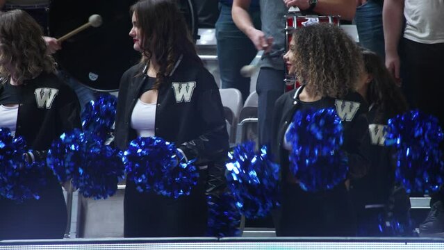 joyful beautiful cheerleaders with pom-poms watching the game and supporting their team, indoors. High quality 4k footage