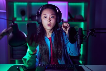 Young asian woman playing video games with smartphone pointing down with fingers showing advertisement, surprised face and open mouth