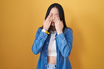 Young asian woman standing over yellow background rubbing eyes for fatigue and headache, sleepy and...