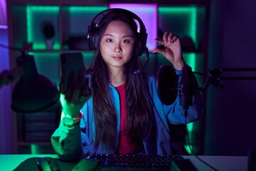 Young asian woman playing video games with smartphone smiling and confident gesturing with hand doing small size sign with fingers looking and the camera. measure concept.