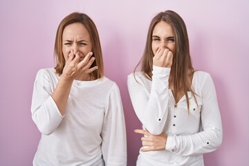 Middle age mother and young daughter standing over pink background smelling something stinky and...
