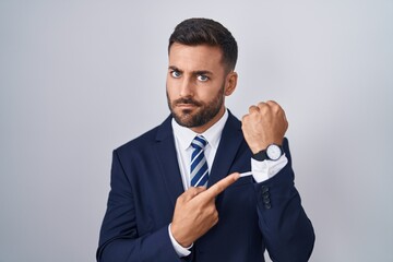 Handsome hispanic man wearing suit and tie in hurry pointing to watch time, impatience, looking at the camera with relaxed expression