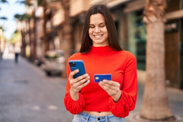 Young beautiful hispanic woman using smartphone and credit card at street