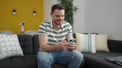 Young caucasian man smiling using smartphone sitting on the sofa at home