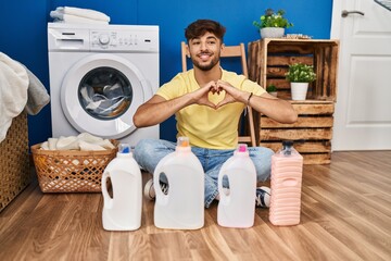 Arab man with beard doing laundry sitting on the floor with detergent bottle smiling in love doing heart symbol shape with hands. romantic concept.