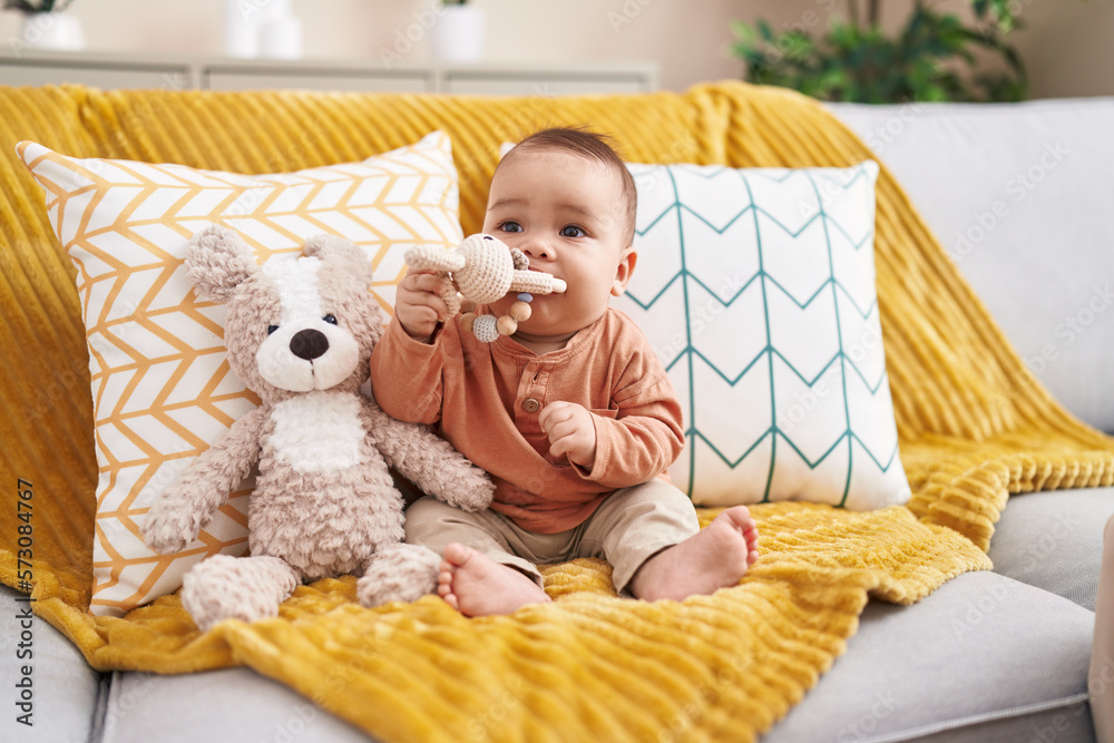 Sticker Adorable hispanic toddler bitting toy sitting on sofa at home