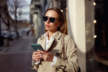 Fashionable woman in sunglasses is standing with phone on city street background and looking at side
