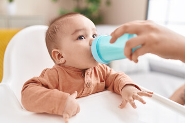 Adorable hispanic toddler drinking water sitting on highchair at home