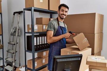 Young hispanic man business worker scanning package using barcode reader machine at storehouse - obrazy, fototapety, plakaty