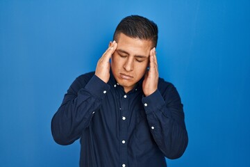 Hispanic young man standing over blue background with hand on head for pain in head because stress. suffering migraine.